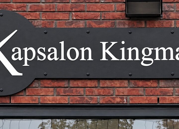 Zwart gevelbord met wit schaarlogo en de naam Kapsalon Kingma, gemonteerd met afstandhouders op een bakstenen gevel