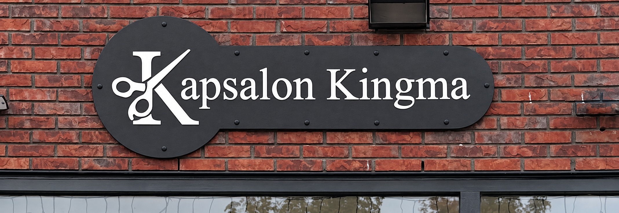 Zwart gevelbord met wit schaarlogo en de naam Kapsalon Kingma, gemonteerd met afstandhouders op een bakstenen gevel